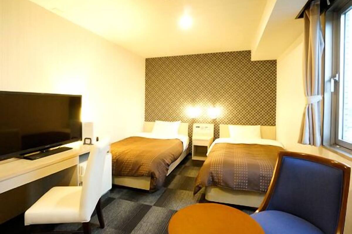 A hotel room with a TV, two beds, a desk, a chair, and a window.