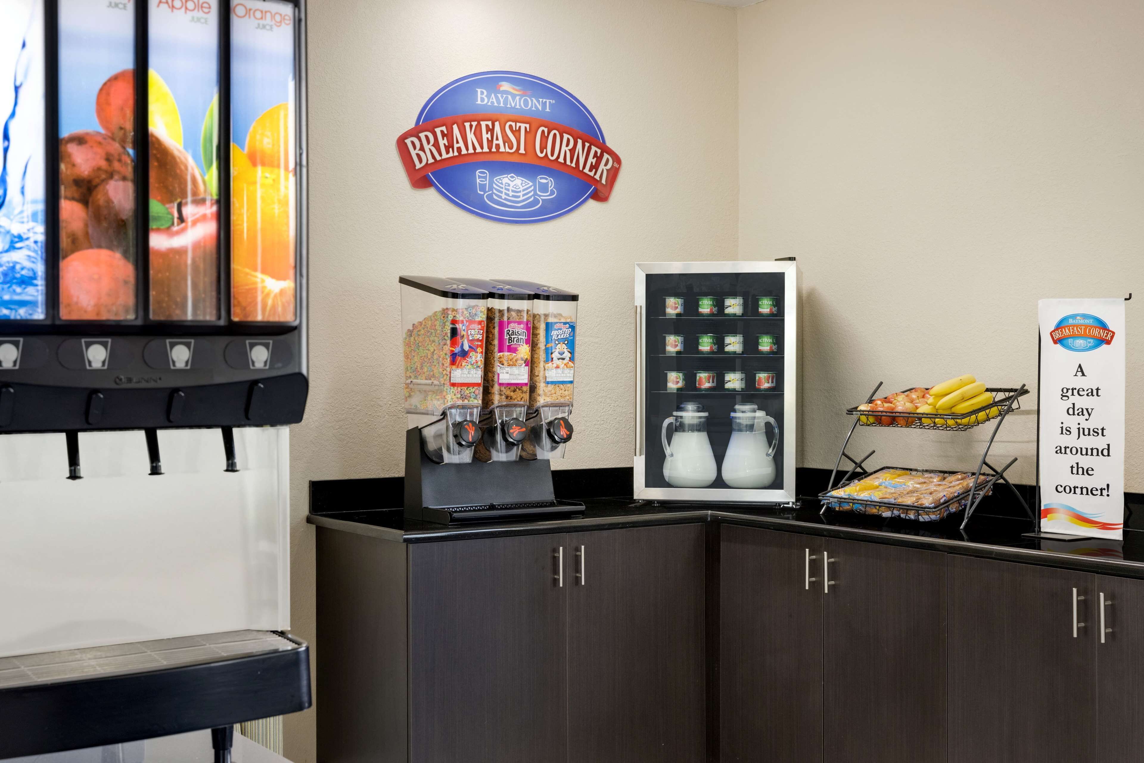 A breakfast station with a Baymont sign, a juice dispenser, a refrigerator with beverages, and a tray of fruit.