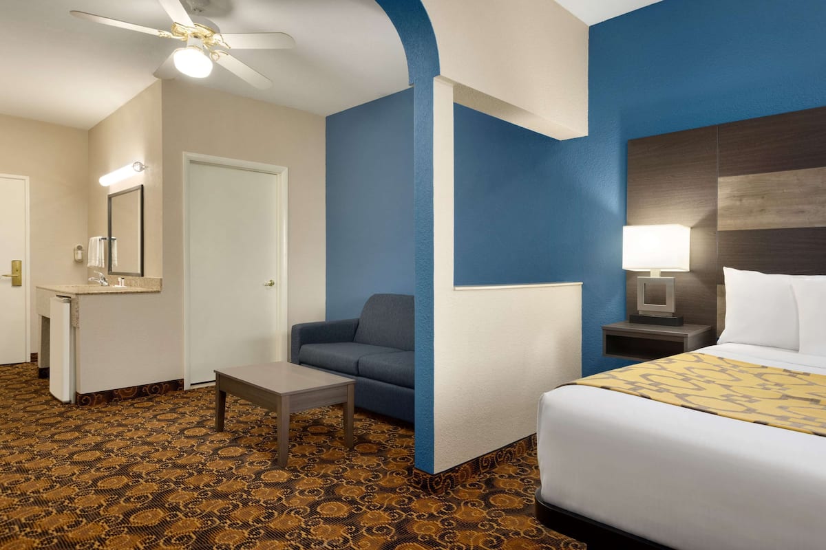 Une chambre d’hôtel comprenant un lit, un canapé, une petite table, une table de chevet, une lampe, un ventilateur de plafond et une salle de bain avec un lavabo et un miroir.