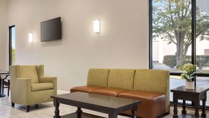 A modern lobby with a sofa, armchair, and a coffee table. A flat-screen TV is mounted on the wall, and there is a window with a view of a tree and building. - Baymont by Wyndham Winchester (Winchester)