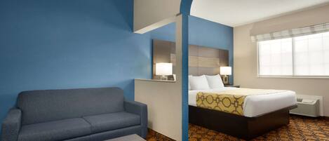 A hotel room with a blue wall, a gray sofa, a bed with a patterned bedspread, a bedside table, and a window with blinds.