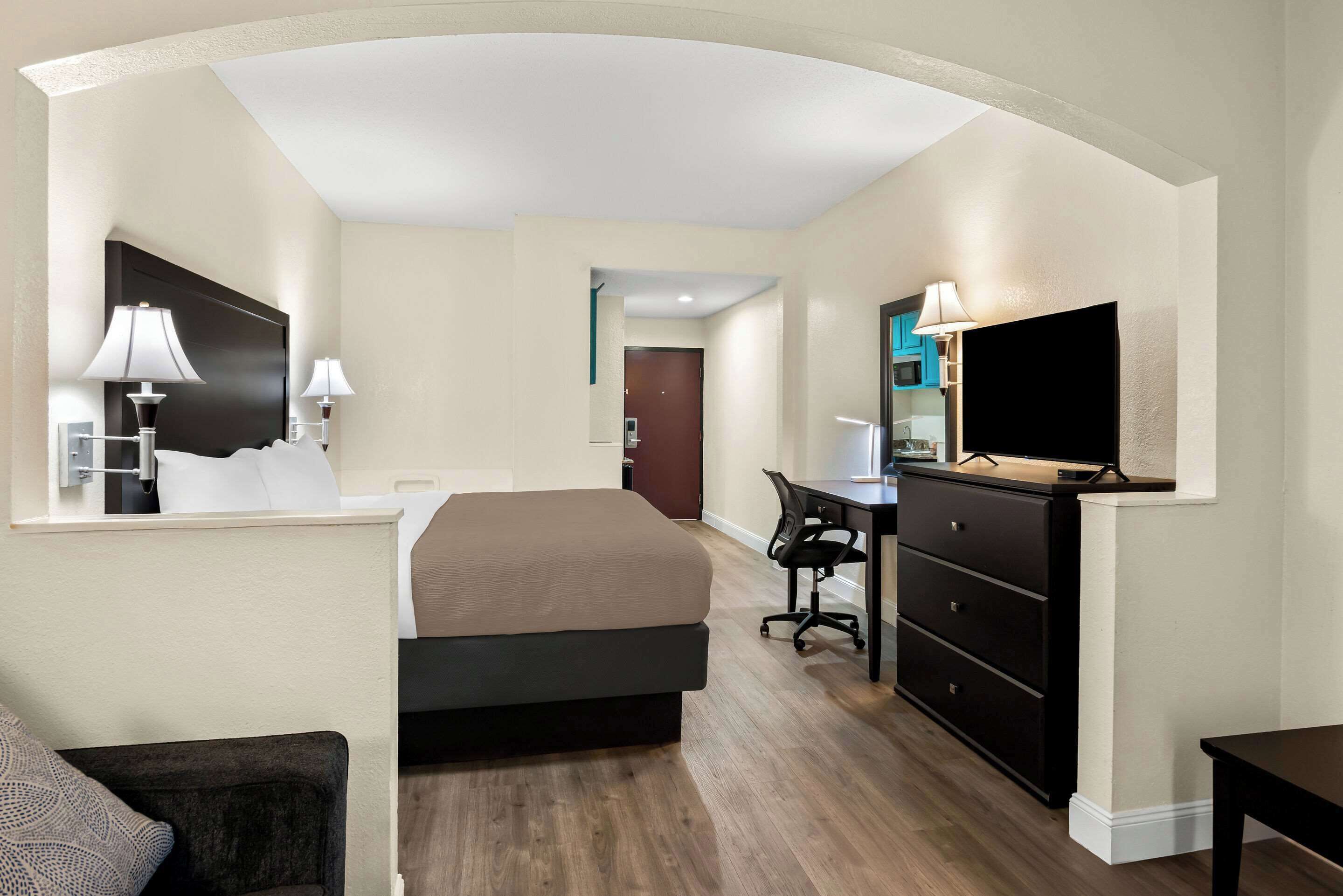 A hotel room with a bed, desk, chair, TV, and a small kitchenette.