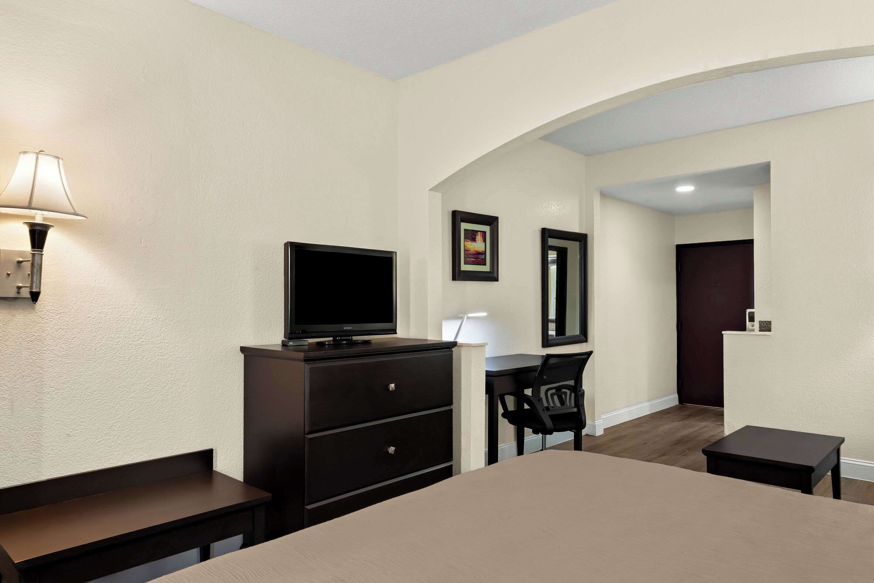 A hotel room with a bed, a desk, a chair, a TV, a lamp, and a door.
