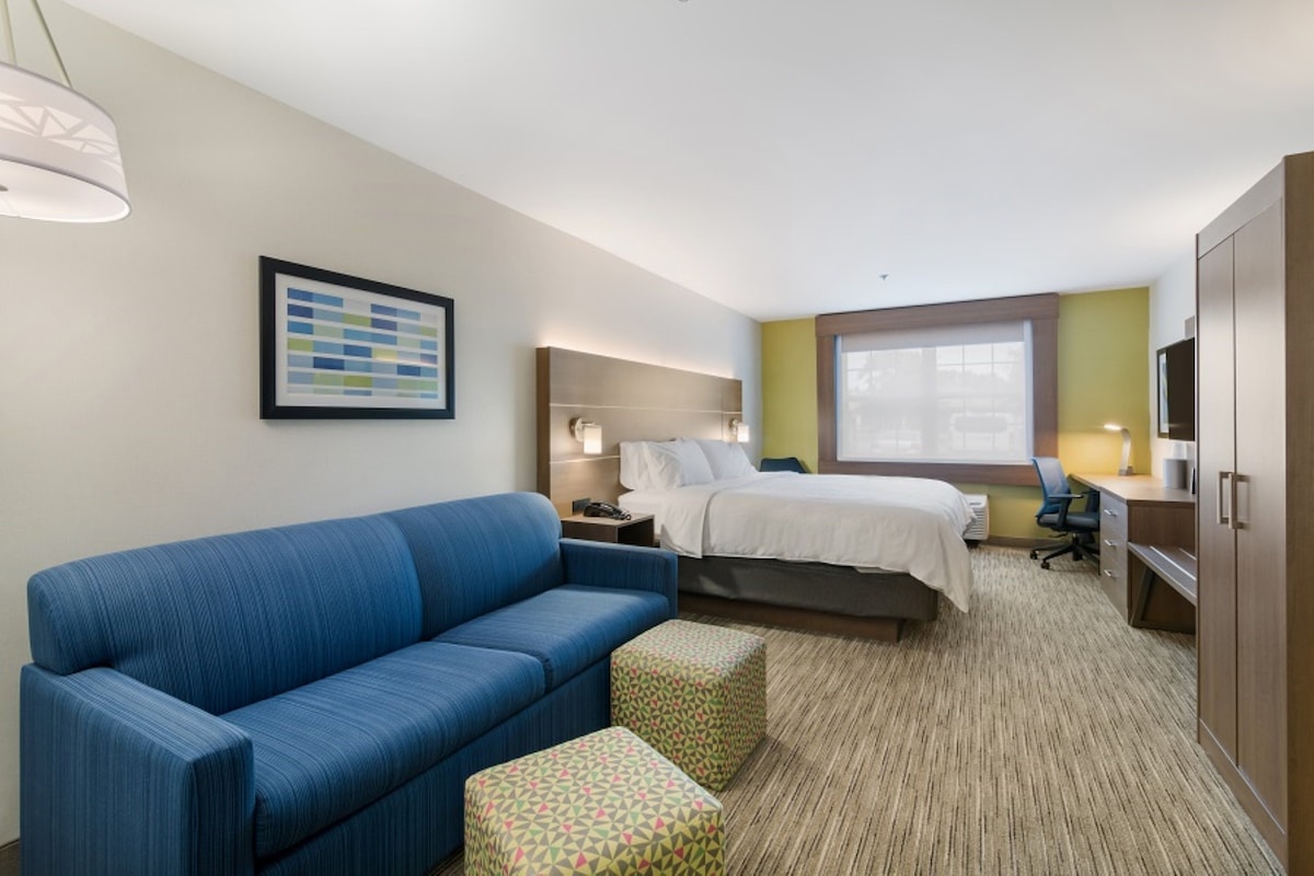 A hotel room with a blue sofa, a bed with white linens, a desk with a chair, a TV, and a framed picture on the wall.