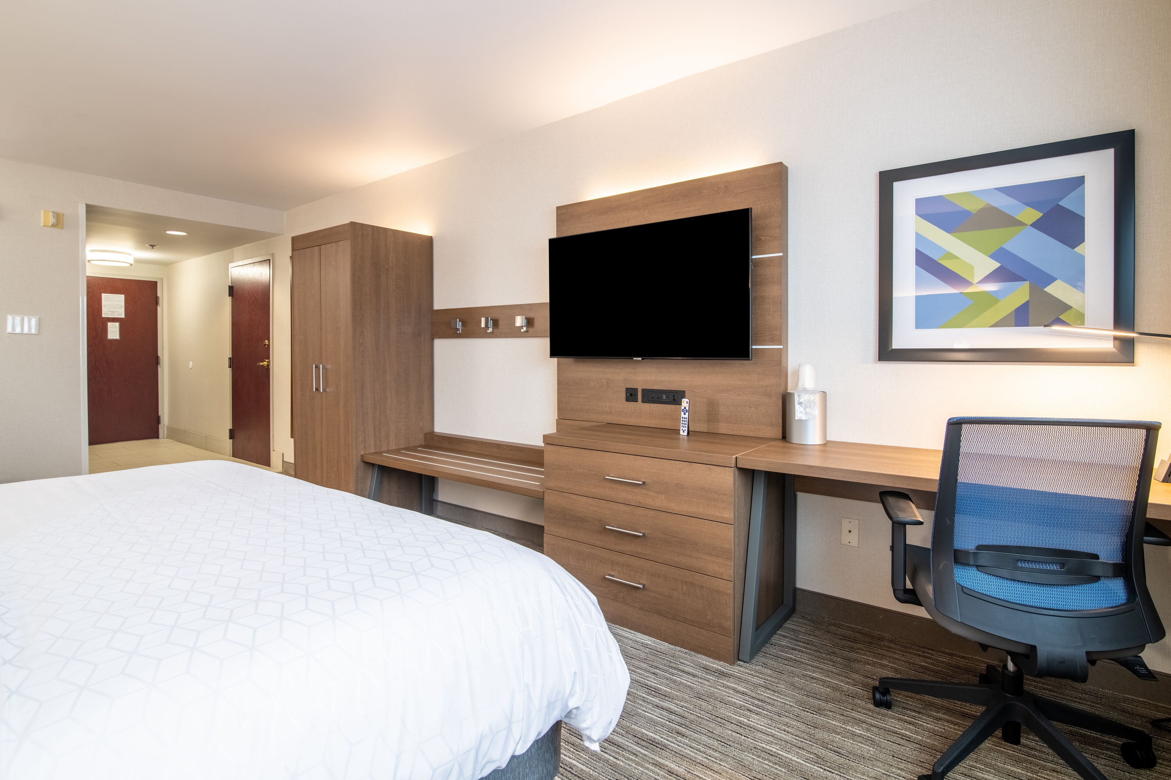 A hotel room with a bed, a desk with a computer, a TV, and a framed picture on the wall.