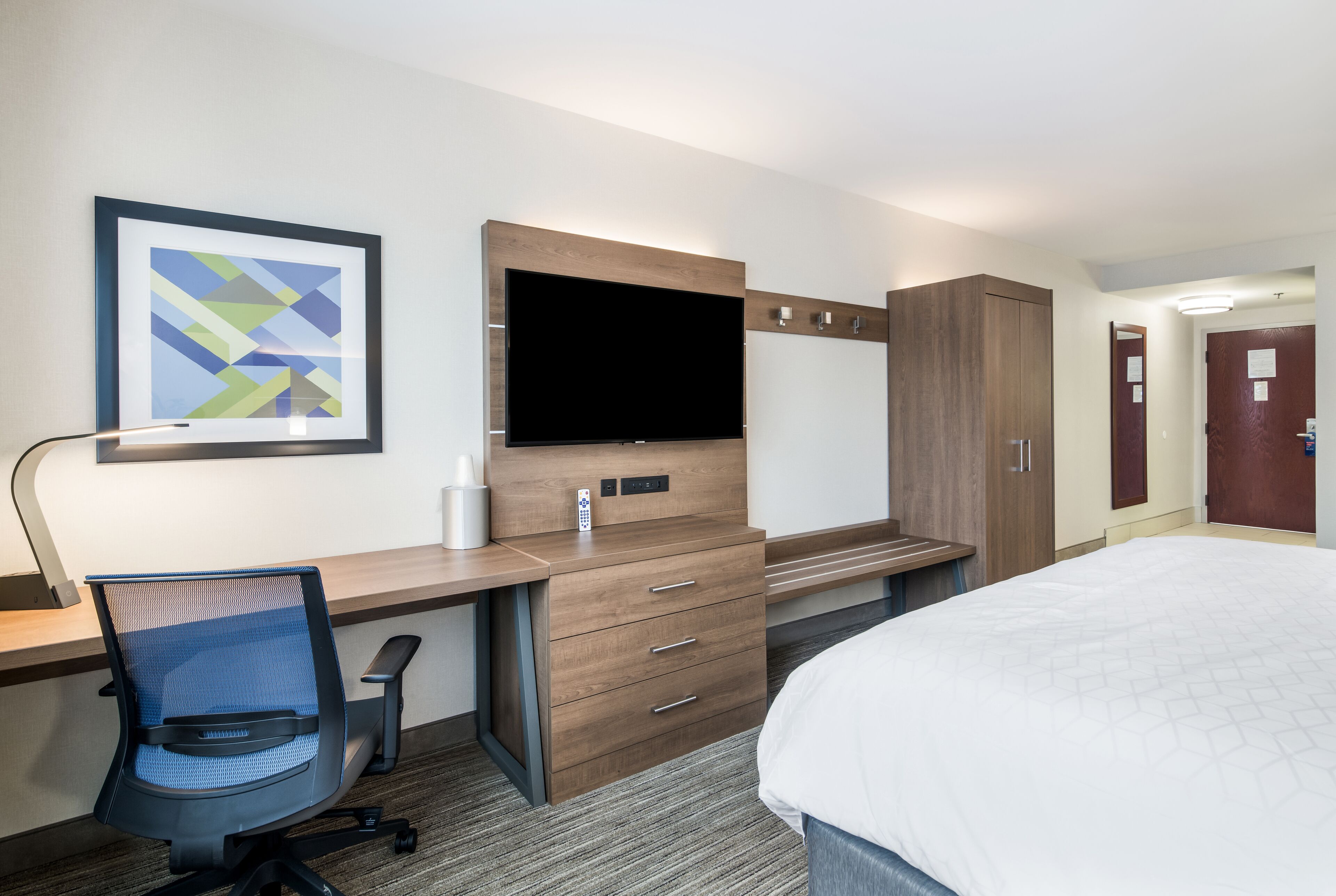 Een hotelkamer met een bed, een bureau met stoel, een televisie, een kledingkast en een lijst aan de muur.