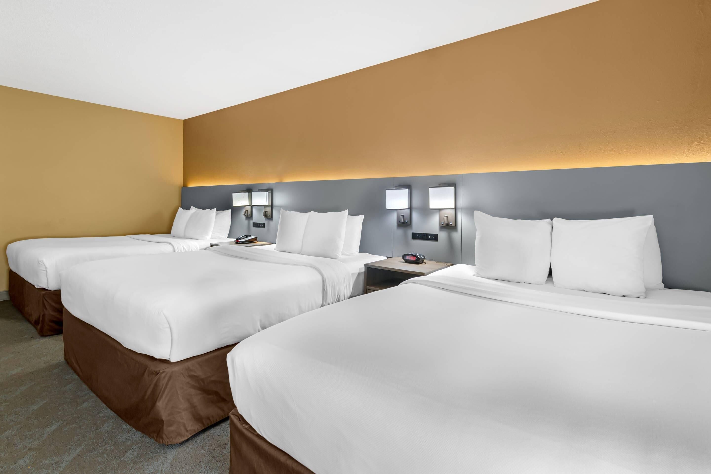 A hotel room with three beds, each with white bedding and brown headboards. There are wall-mounted light fixtures above each bed.