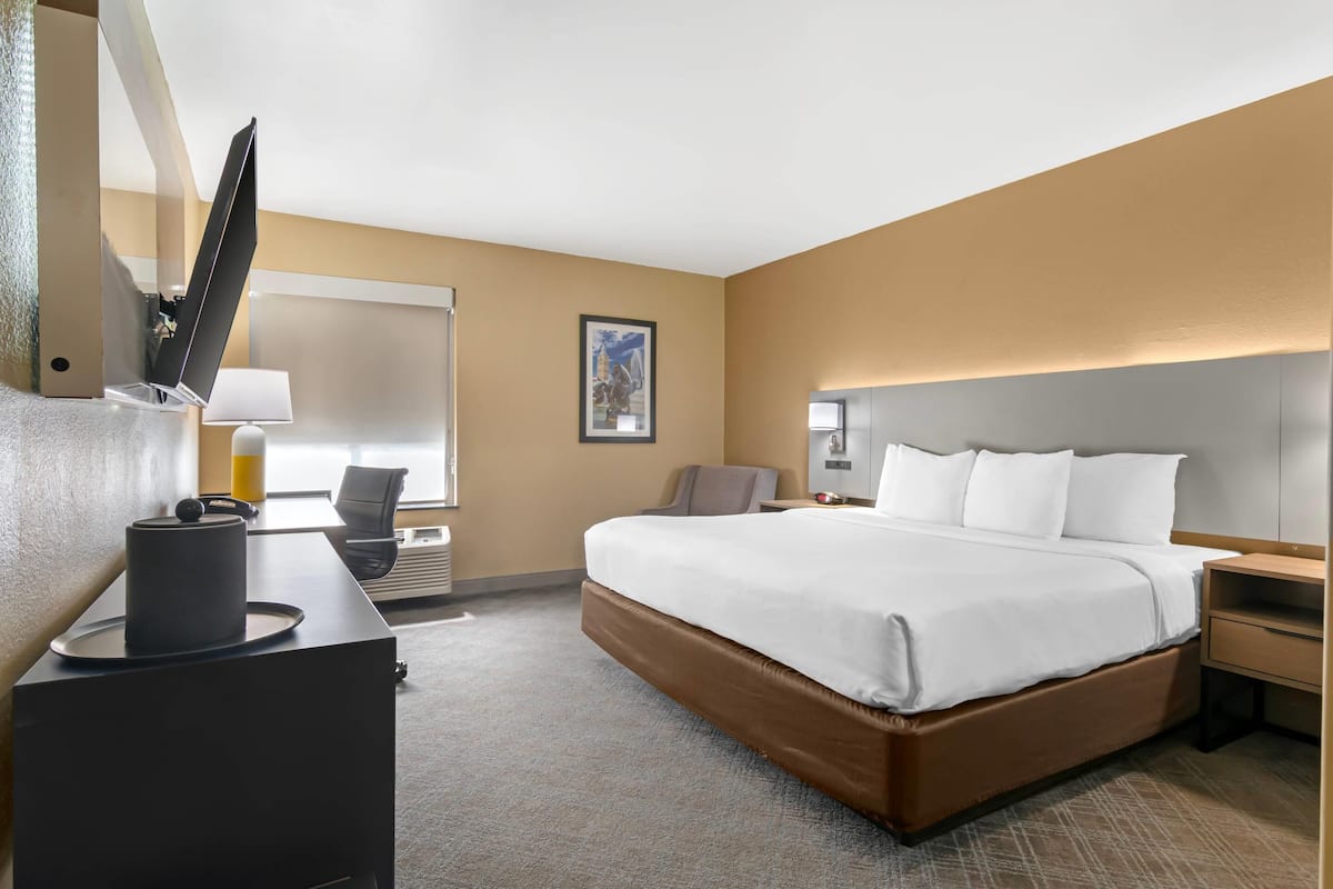 Quarto de hotel com uma cama grande, uma escrivaninha com abajur, uma cadeira e uma televisão.