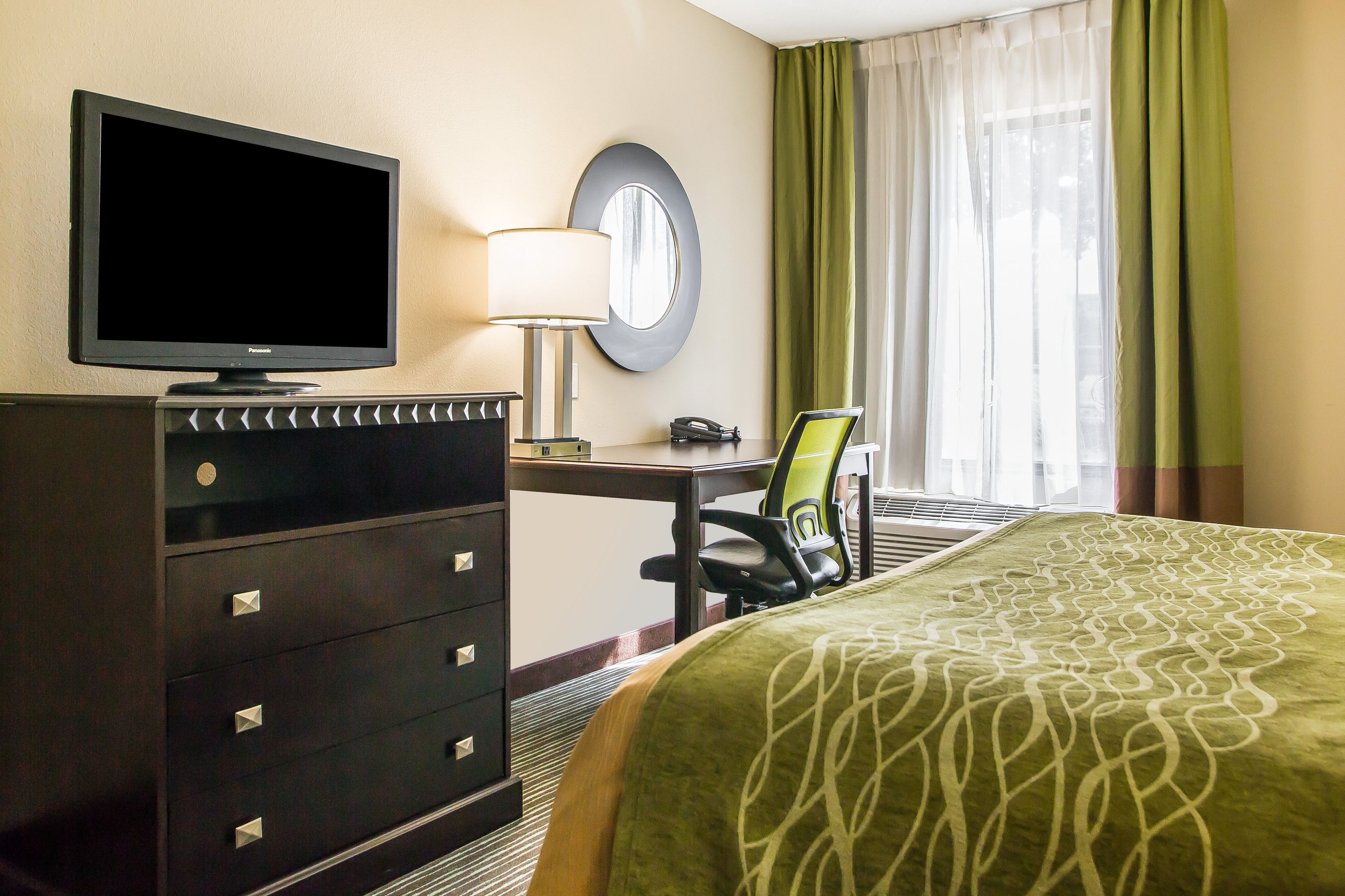 A hotel room with a television, a desk with a lamp, a chair, a bed with a patterned bedspread, and a window with curtains.