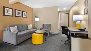 A hotel room with a grey sofa, a small round table, a yellow ottoman, a grey armchair, a desk with a lamp, and framed pictures on the wall. - Comfort Inn & Suites Kansas City - Northeast (Kansas City)
