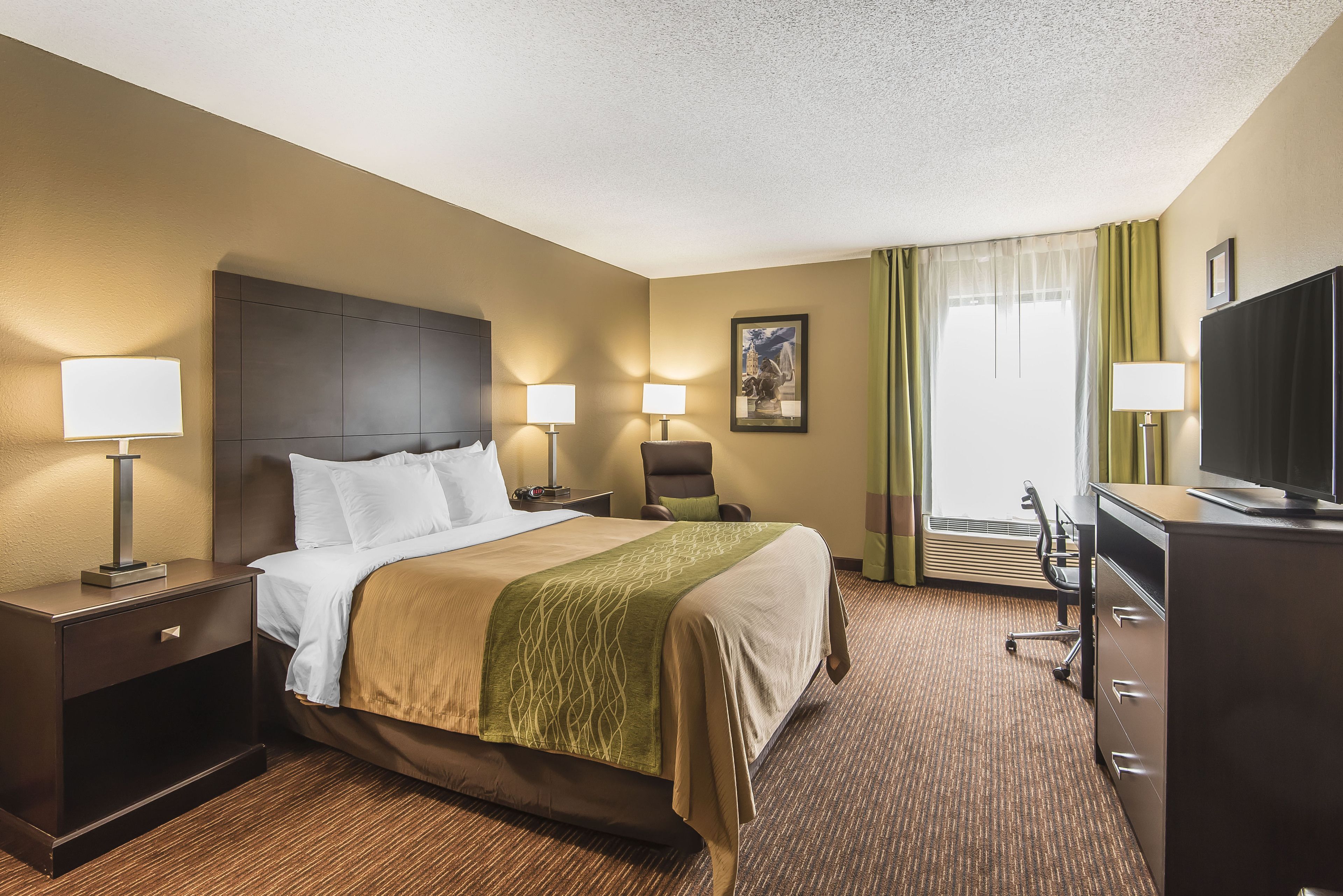 A hotel room with a bed, two bedside tables with lamps, a desk with a chair, a television, and a window with curtains.