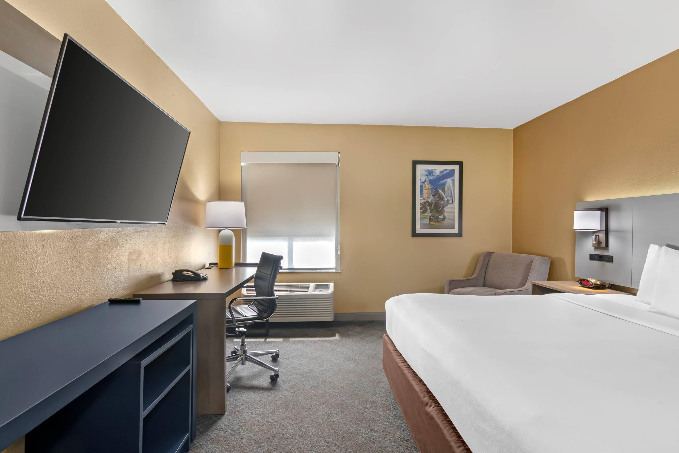 A hotel room with a bed, a desk with a chair, a television, a window with blinds, and a framed picture on the wall.