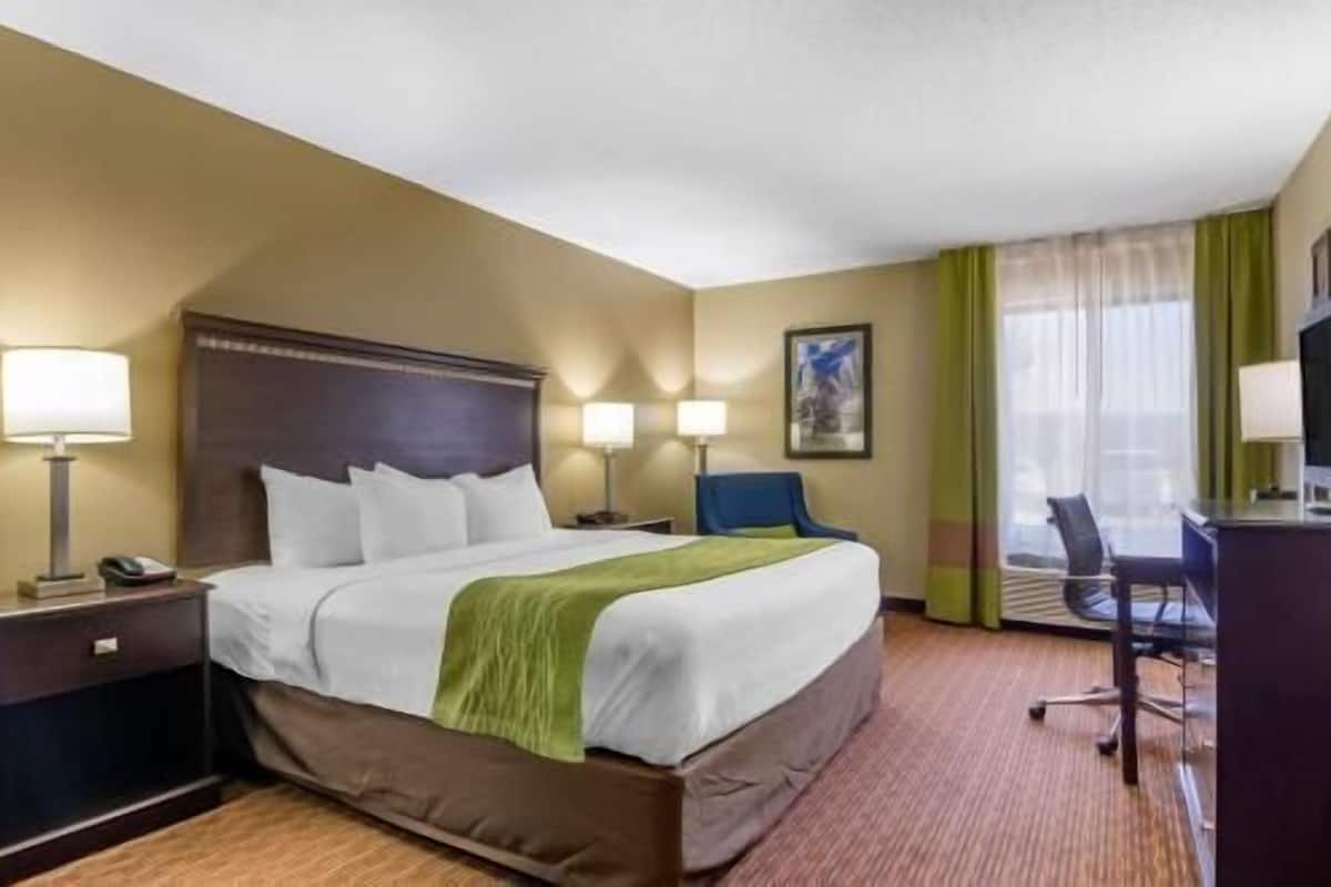 Quarto de hotel com cama, duas luminárias de cabeceira, escrivaninha com cadeira e janela com cortinas.