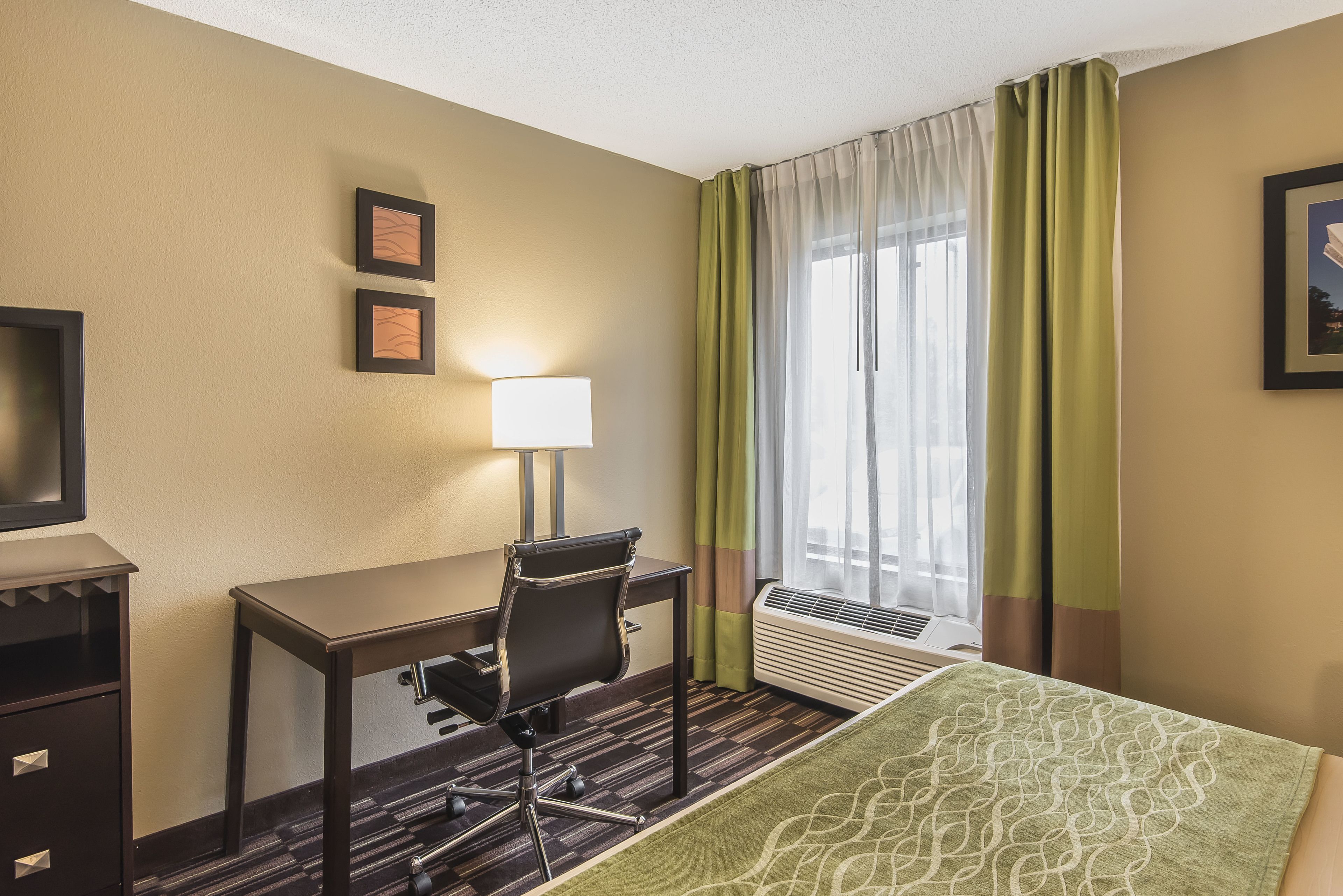 A hotel room with a desk, chair, and a bed with a green patterned bedspread. There is a window with sheer curtains and a wall-mounted air conditioner.
