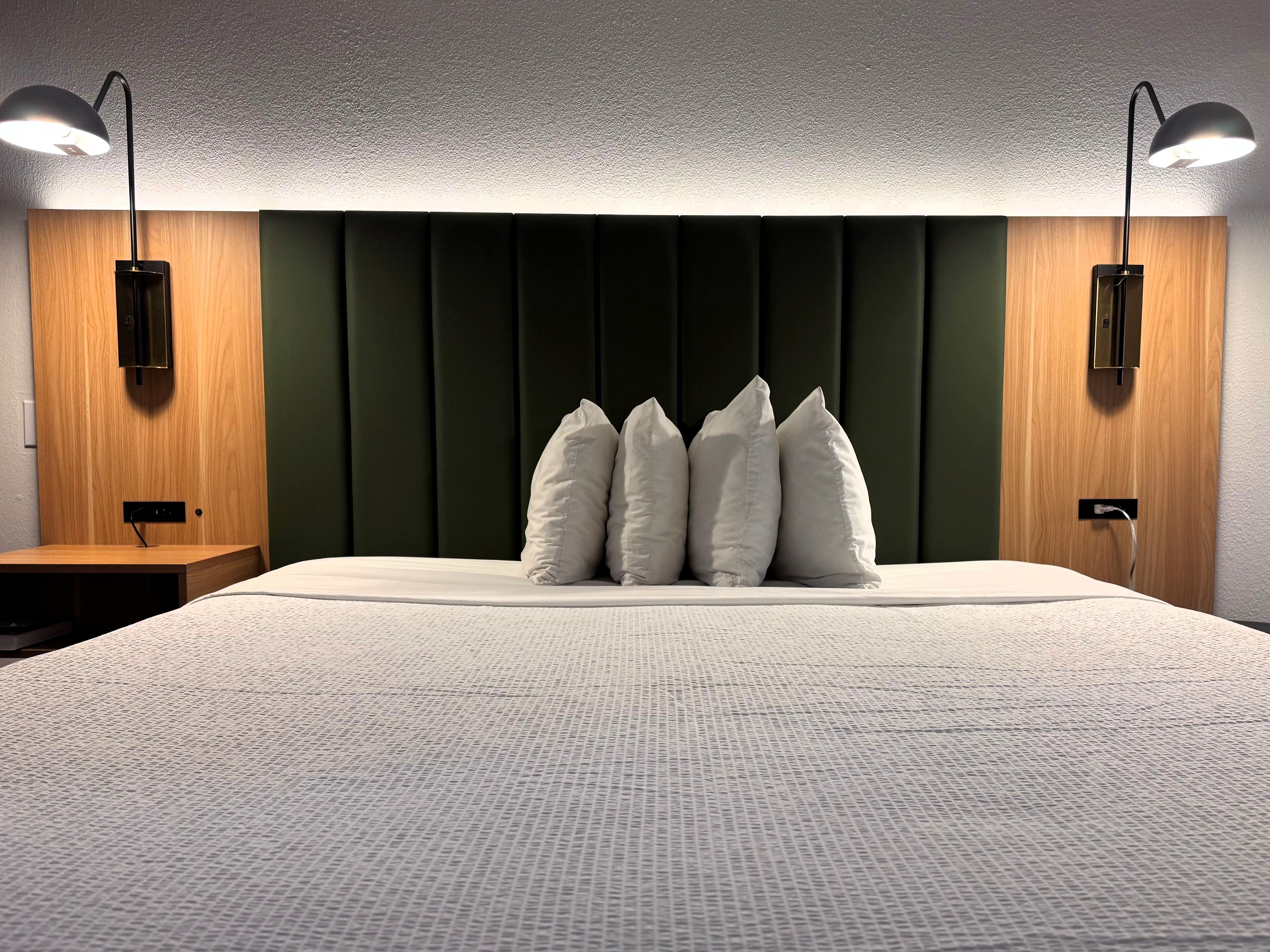 A bed with a textured white cover, a dark green upholstered headboard, and three white pillows. There are two wall-mounted lamps on either side of the bed.
