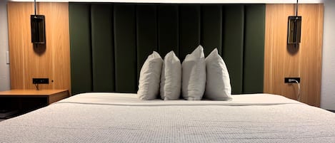 A bed with a textured white cover, a dark green upholstered headboard, and three white pillows. There are two wall-mounted lamps on either side of the bed.