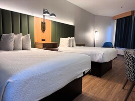 Habitación de hotel con dos camas, cabecero de madera, mesita de noche, una silla y un ventanal grande con cortinas.