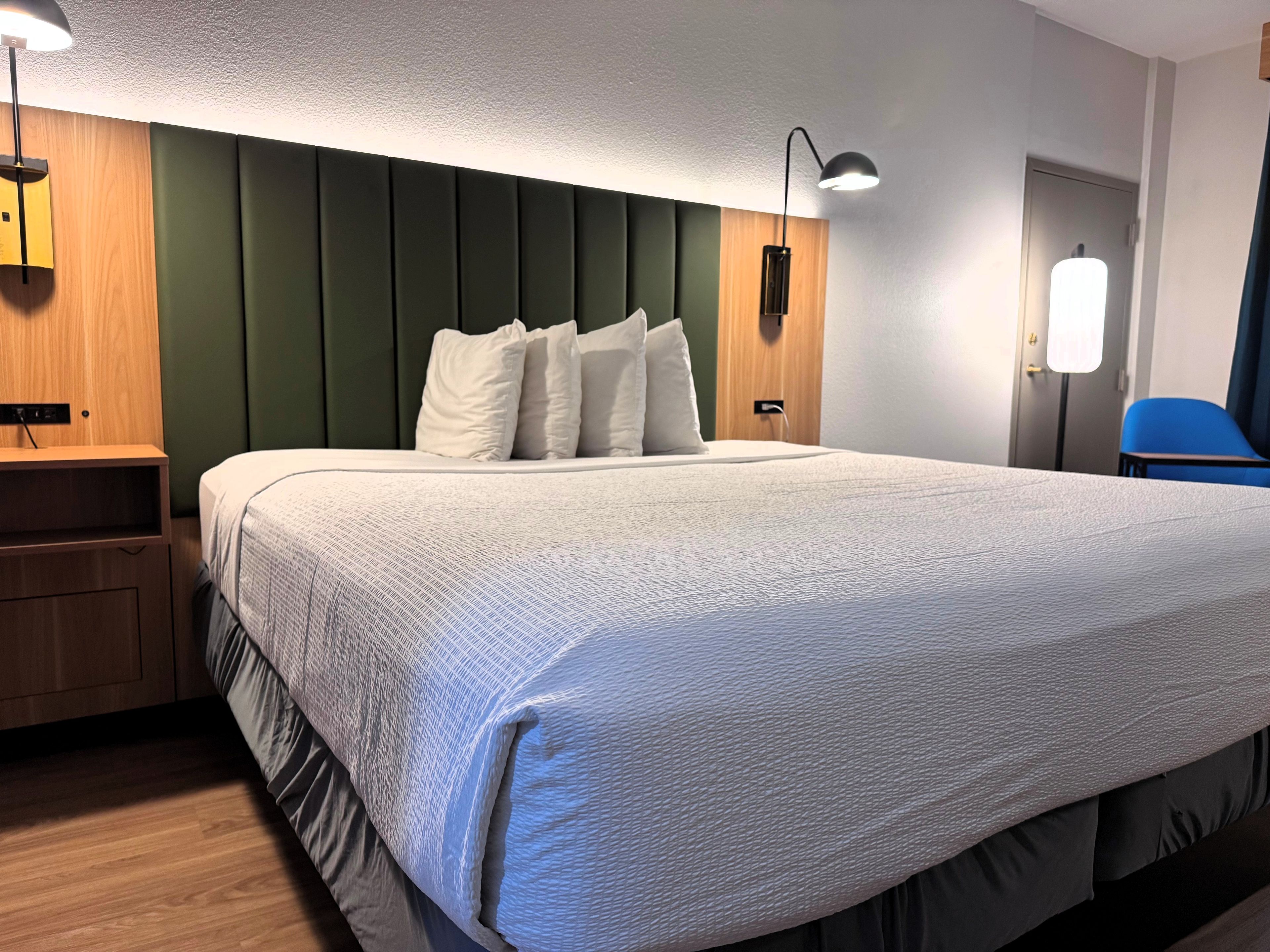 A neatly made bed with a white comforter and pillows, a wooden nightstand with a lamp, and a blue chair in a room with green walls and a wooden door.