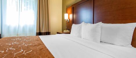 A hotel room with a large bed, a wooden headboard, a bedside table with a lamp, and a window with curtains.