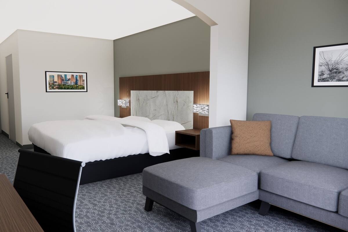 A modern hotel room with a bed, a grey sofa, a wooden desk, and a cityscape picture on the wall.