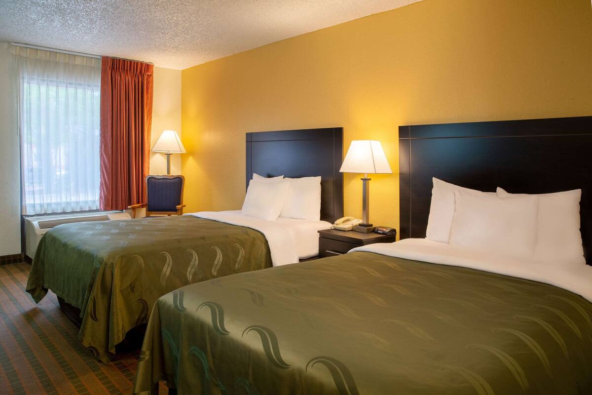 A hotel room with two beds, each with a headboard, a bedside table with a lamp, and a window with curtains.