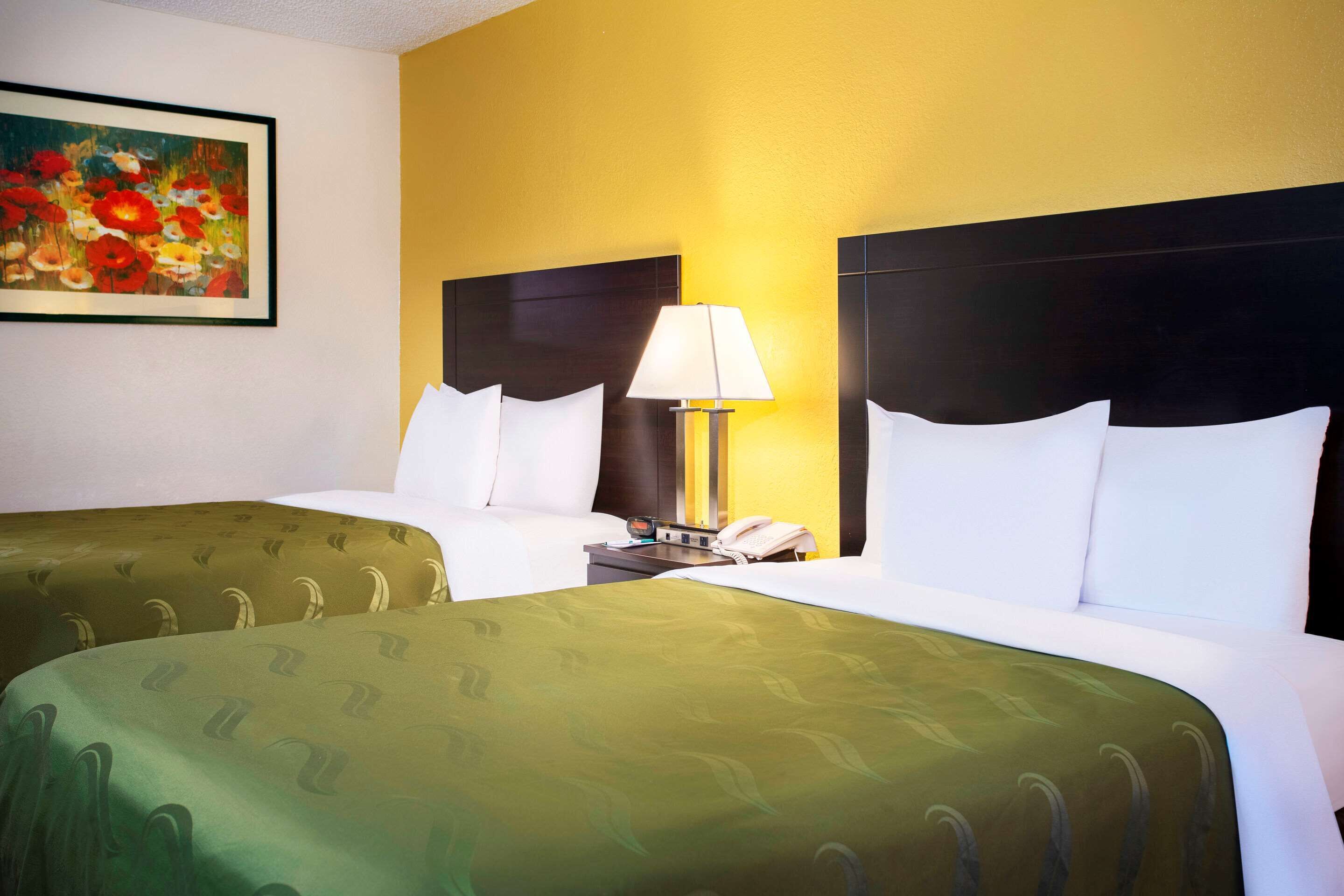 Quarto de hotel com duas camas, uma mesa de cabeceira com abajur, um quadro na parede e um telefone.