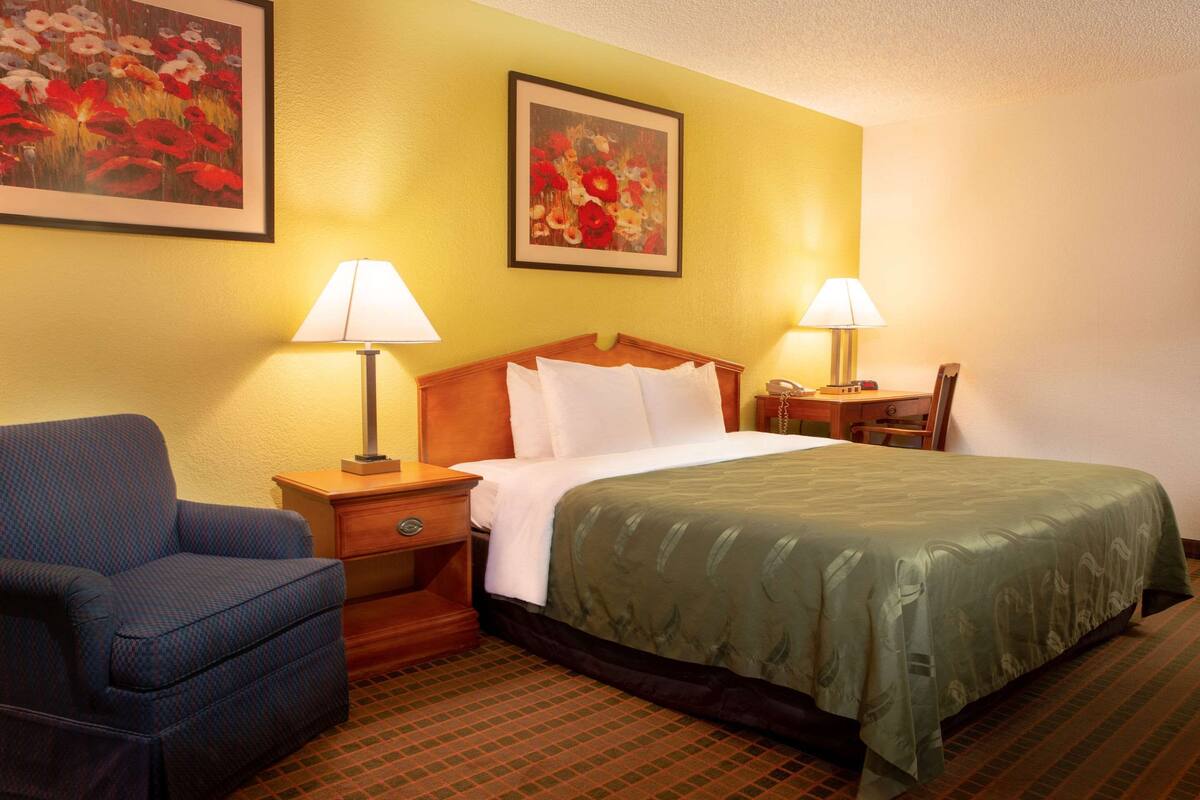 A hotel room with a bed, two bedside lamps, a chair, and two framed floral paintings on the wall.