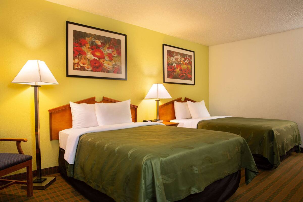 A hotel room with two beds, each with a green bedspread, two bedside lamps, and two framed floral paintings on the wall.