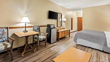 Habitación de hotel con una cama, un escritorio con silla, un refrigerador pequeño, televisor y lámpara.