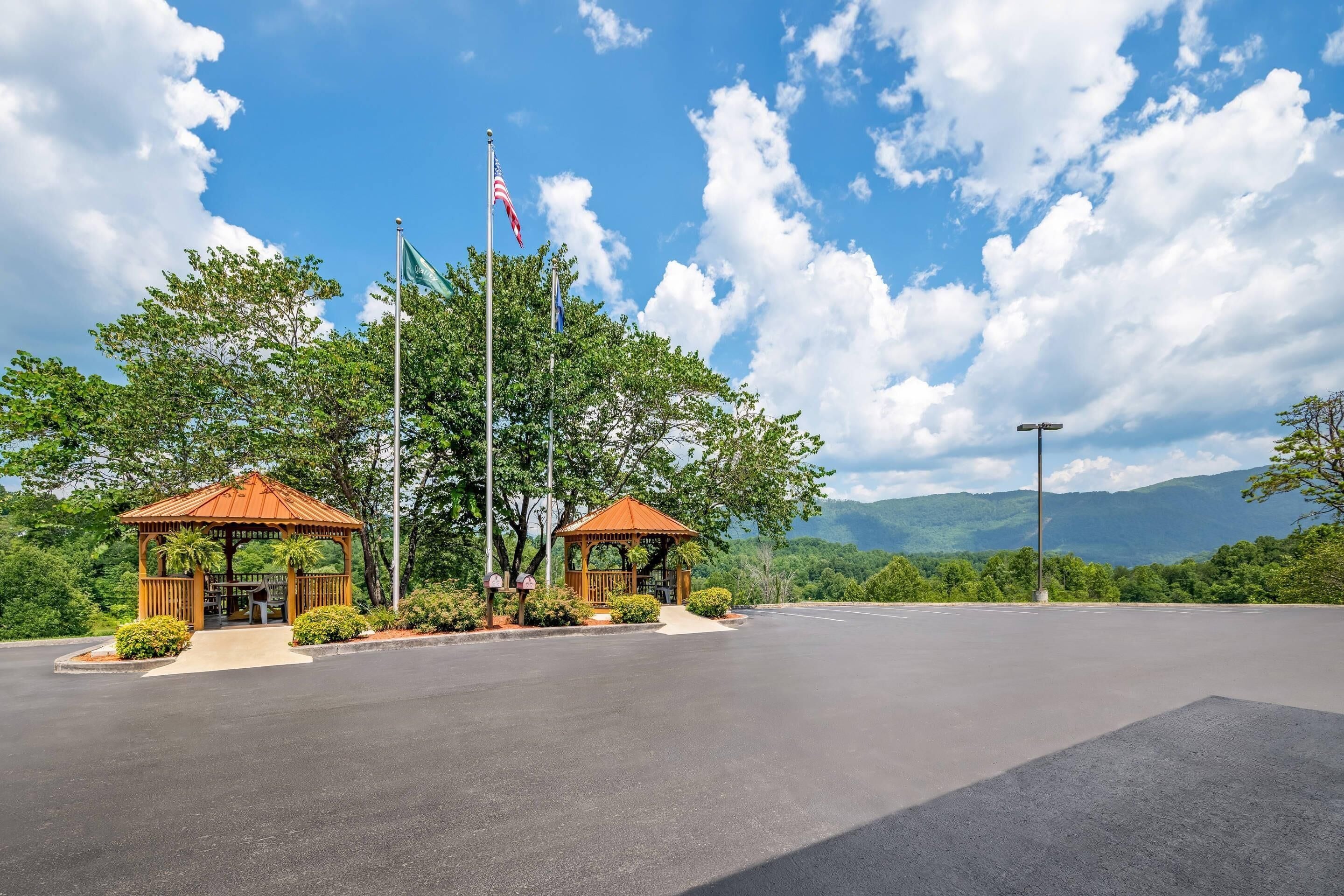 A gazebo with a red roof and two flagpoles, one with the American flag, in a parking lot with a mountain view.