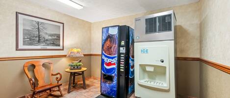 Un vestíbulo de hotel con una mecedora de madera, un refrigerador Pepsi, un dispensador de hielo, un cuadro en la pared y una pequeña mesa con una lámpara.
