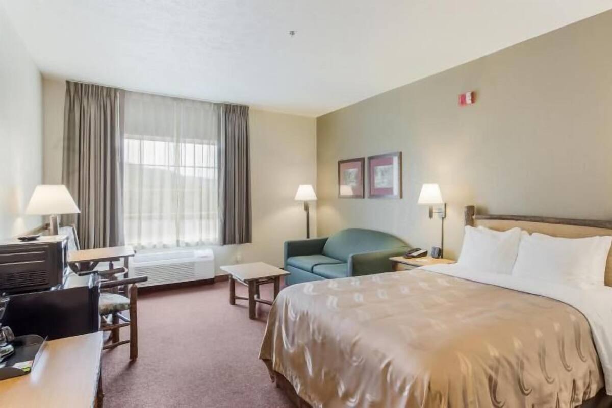 Quarto de hotel com cama, duas mesas de cabeceira, escrivaninha, poltrona, sofá, janela com cortinas e duas luminárias fixadas na parede.