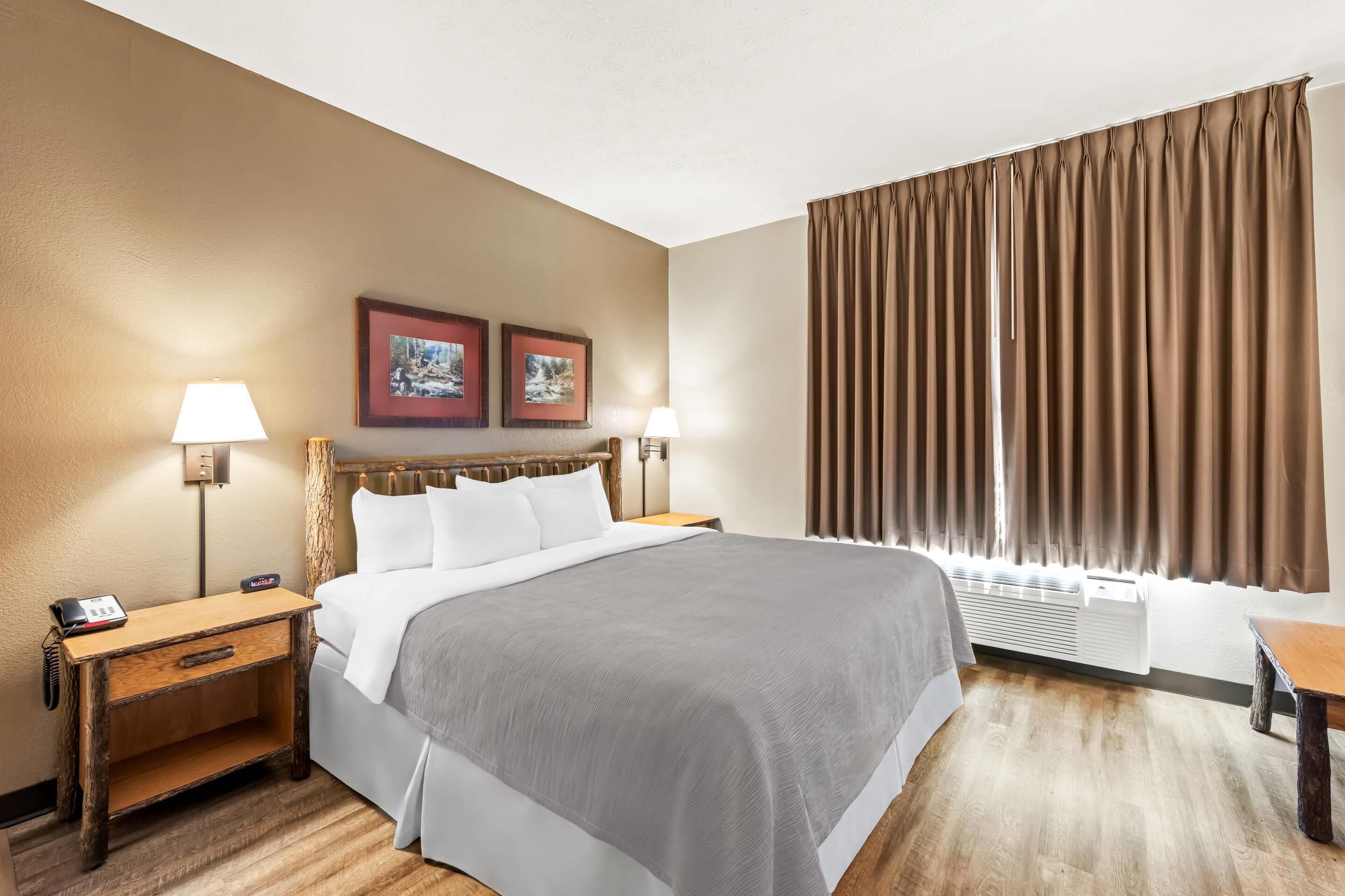 Une chambre d’hôtel avec un grand lit, deux tables de chevet avec des lampes, une petite table en bois et une grande fenêtre avec des rideaux.