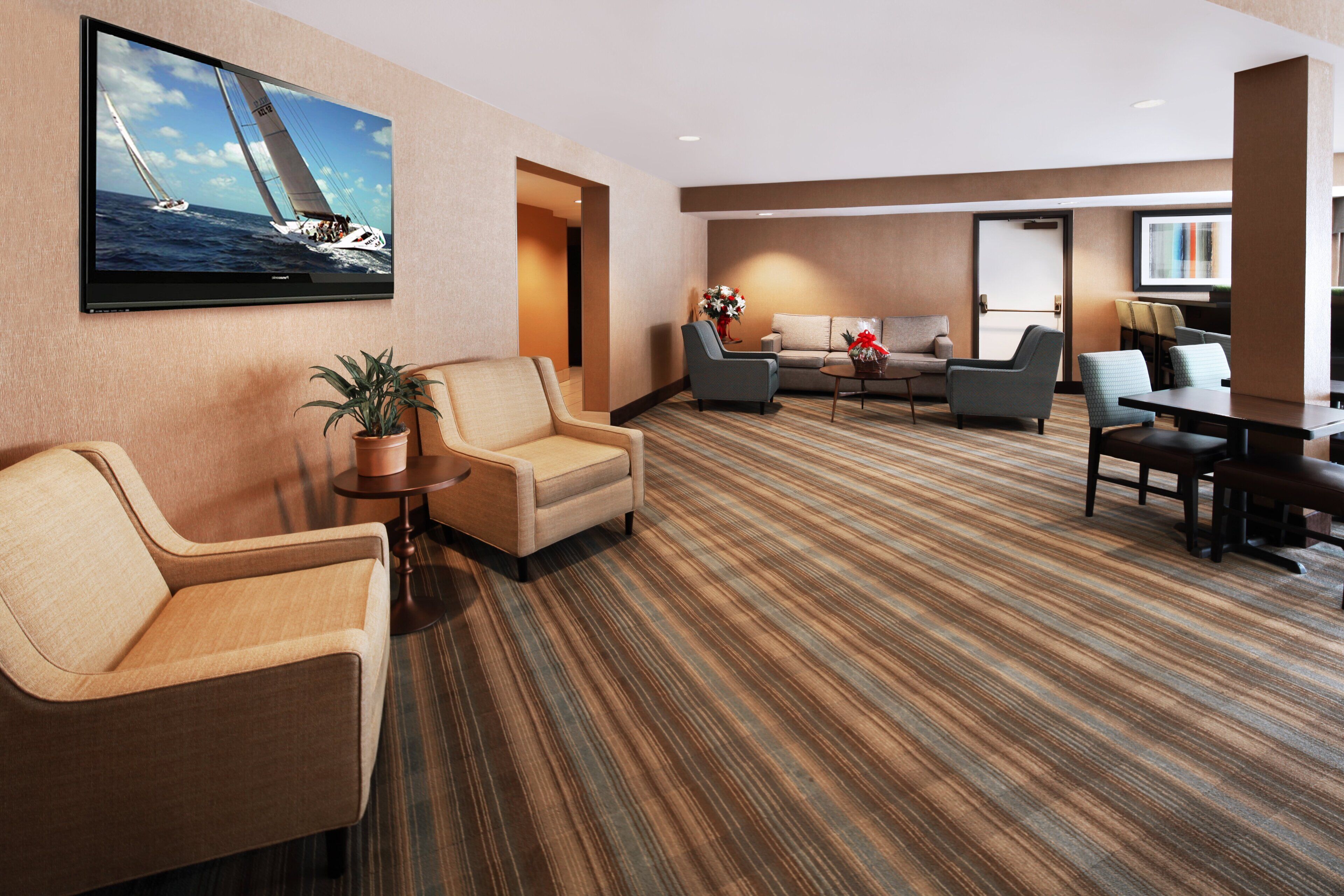 Un hall de hotel con un televisor de pantalla plana grande mostrando un velero, una zona de estar con sofás y sillones, y una alfombra a rayas.