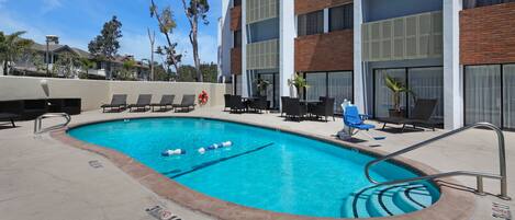 A swimming pool with lounge chairs and a lifebuoy, surrounded by a building with brick and white walls, and trees in the background.