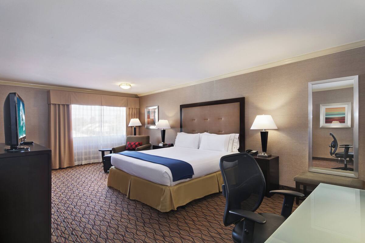A hotel room with a large bed, two bedside tables with lamps, a desk with a chair, a television, and a large mirror on the wall.