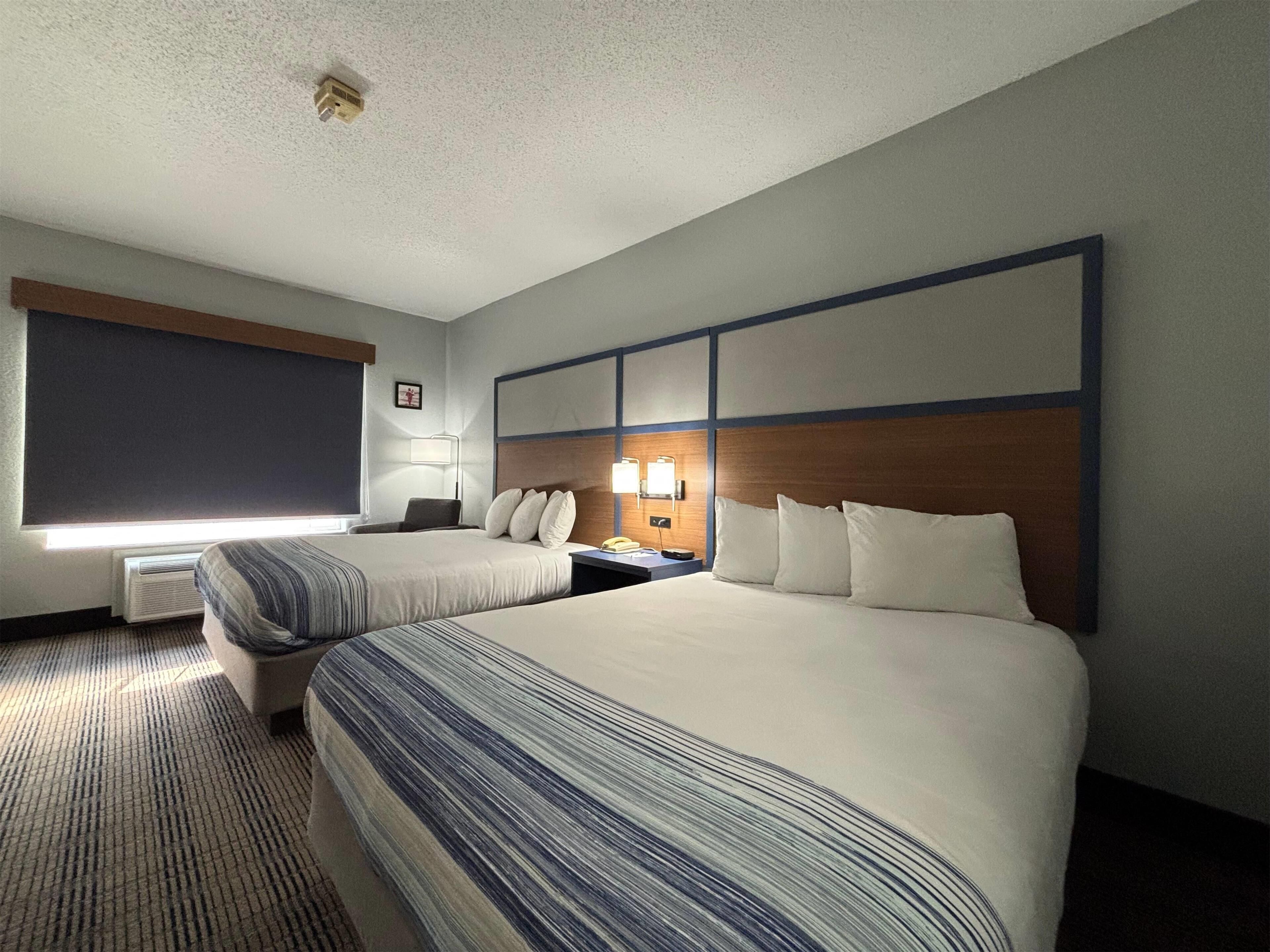 A hotel room with two beds, a desk, a chair, a lamp, and a window with blinds.