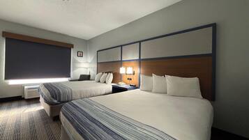 A hotel room with two beds, a desk, a chair, a lamp, and a window with blinds.