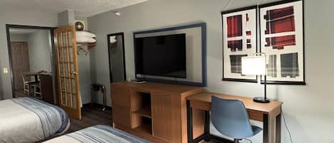 A hotel room with two beds, a desk with a chair, a television, a mirror, and a framed picture on the wall.