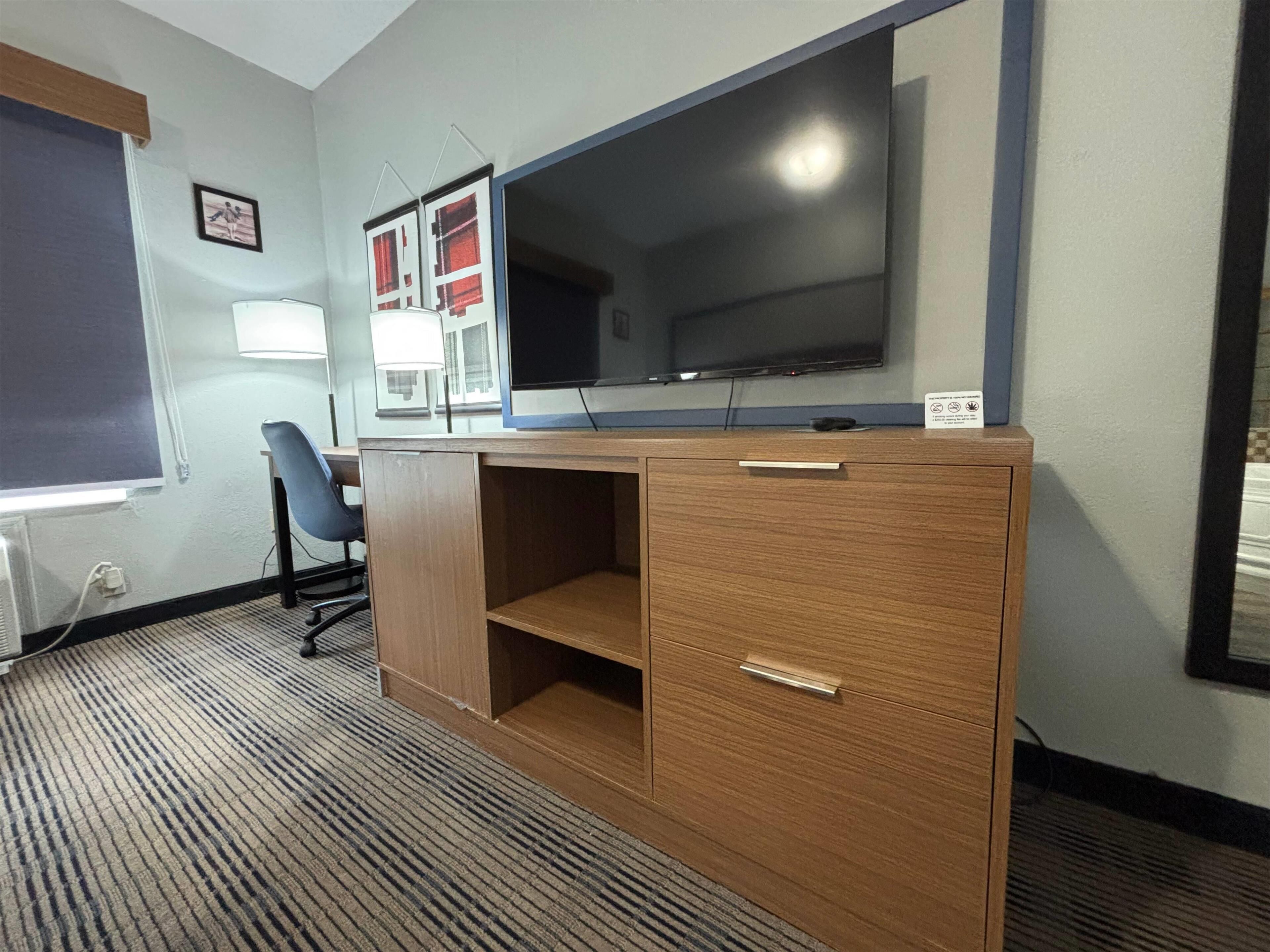 A hotel room with a wooden desk, a flat-screen TV, a chair, and a window.