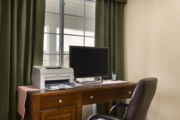 A desk with a printer, monitor, and chair in a room with a window and green curtains.