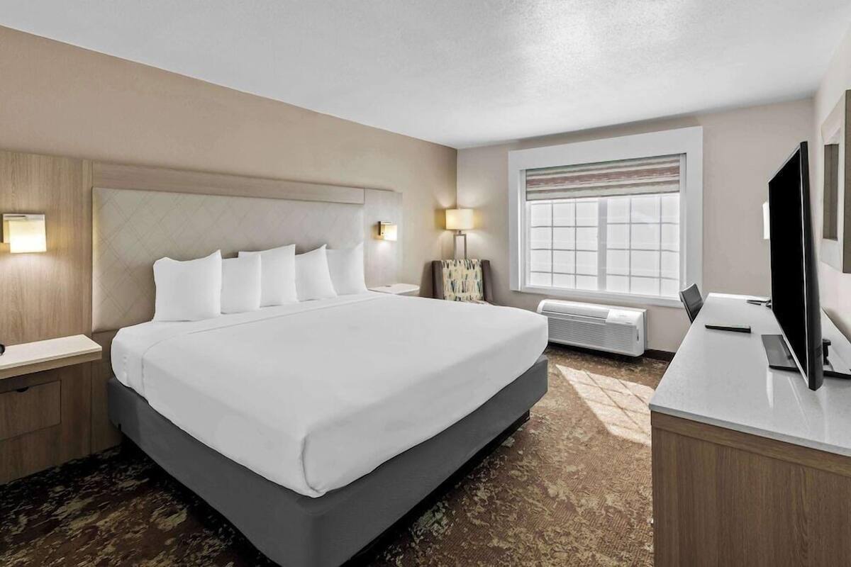 A hotel room with a large bed, bedside tables, a desk with a TV, a window with blinds, and a wall-mounted air conditioning unit.