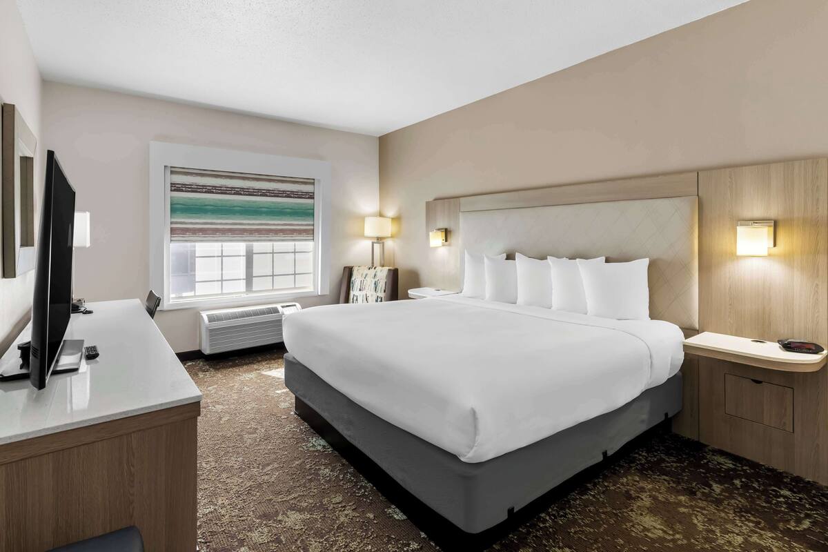 A hotel room with a large bed, a desk with a flat-screen TV, a window with a view, and wall-mounted lamps.