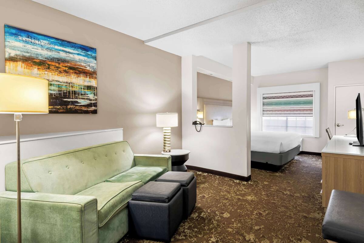 A hotel room with a green sofa, a small table, a lamp, a bed, a TV, and a painting on the wall.