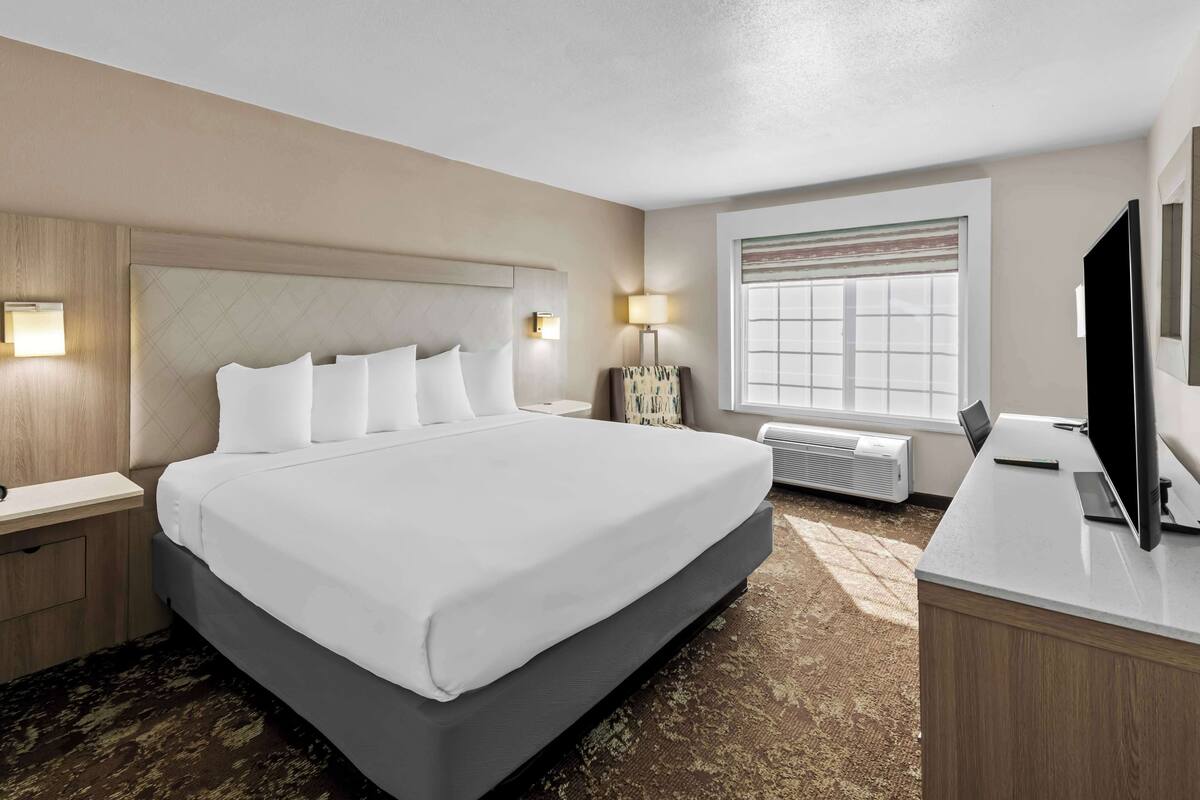 A hotel room with a large bed, bedside tables, a desk with a TV, and a window with blinds.