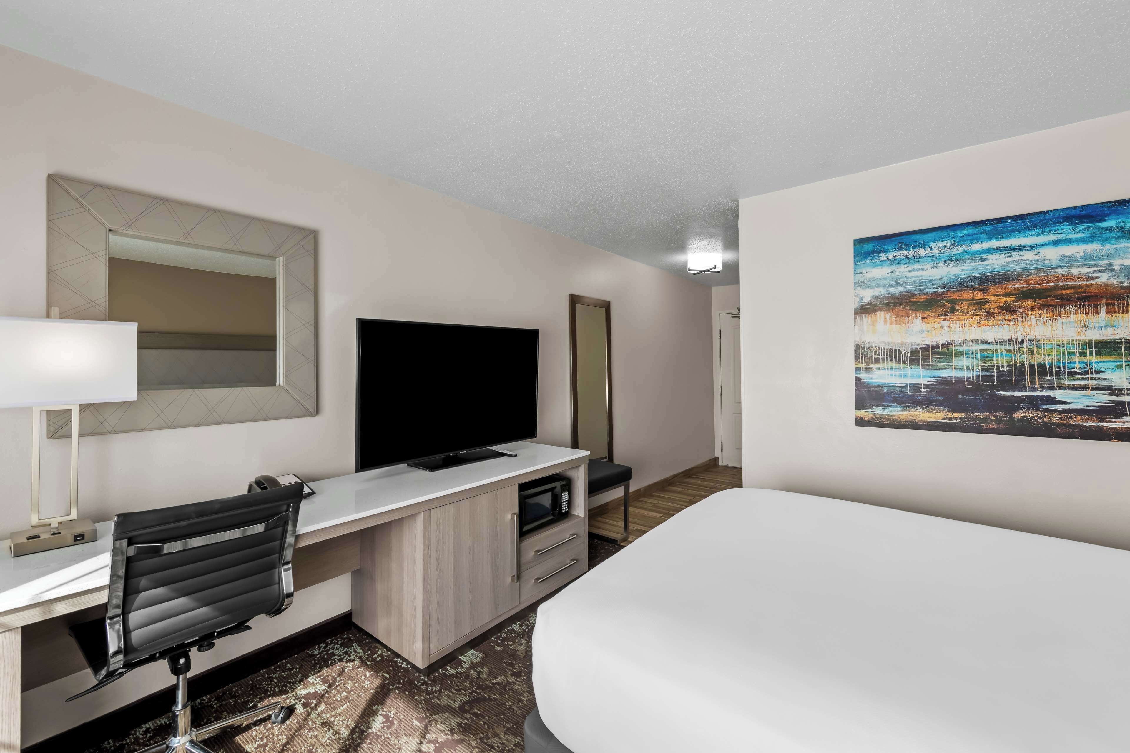 A hotel room with a bed, a desk with a flat-screen TV, a mirror, and a painting on the wall.
