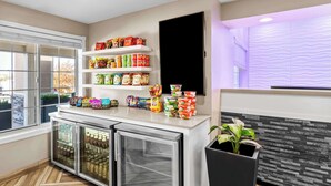 A hotel room with a minibar, shelves with snacks and beverages, a refrigerator, a potted plant, and a window with a view. - Best Western Worlds of Fun Inn & Suites (Kansas City)