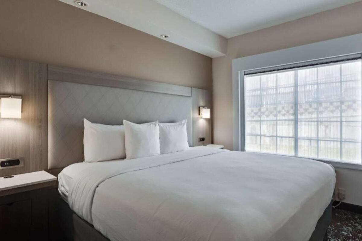 A hotel room with a large bed, two bedside tables with lamps, a window with blinds, and a wall-mounted control panel.