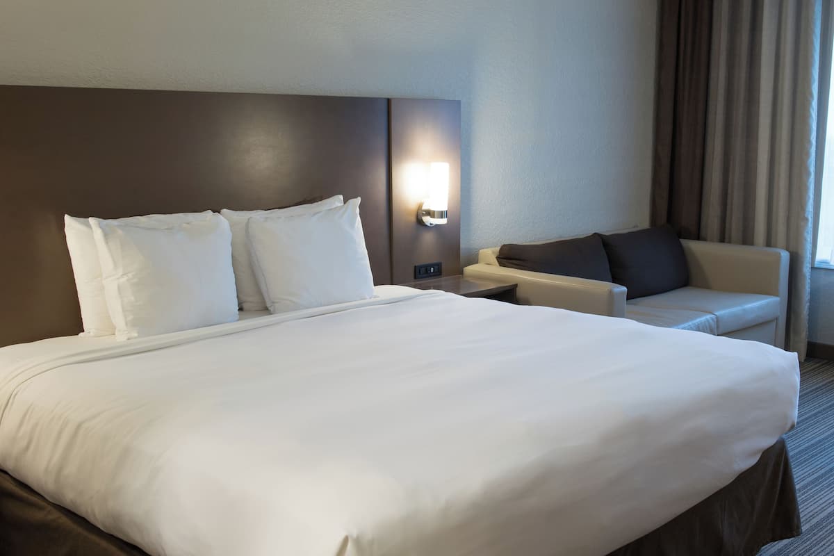 A hotel room with a large bed, a sofa, a nightstand, and a wall-mounted light.