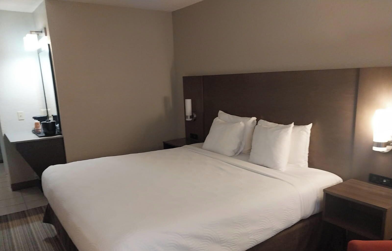 A hotel room with a large bed, a bedside table, a nightstand, a mirror, and a wall-mounted light.